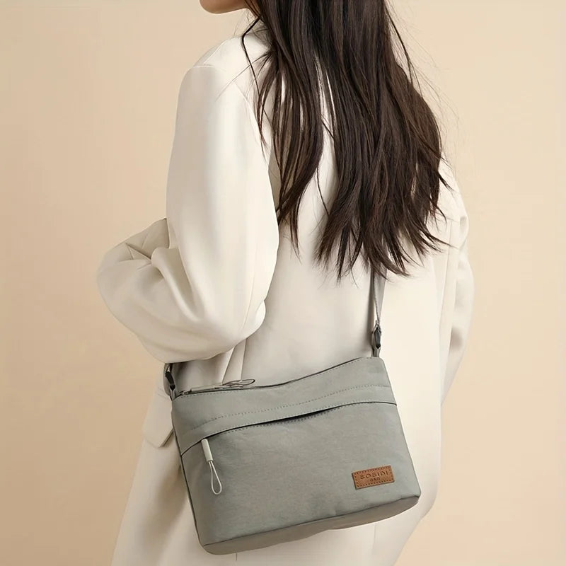 A light gray nylon crossbody shoulder bag with a leather tag is carried by a person wearing a cream-colored coat.