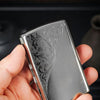 A hand holds a compact silver metal cigarette case featuring intricate embossed scrollwork.