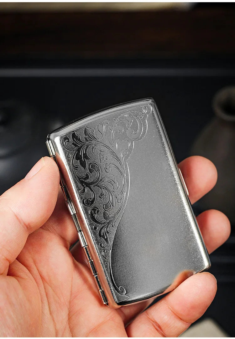 A hand holds a compact silver metal cigarette case featuring intricate embossed scrollwork.