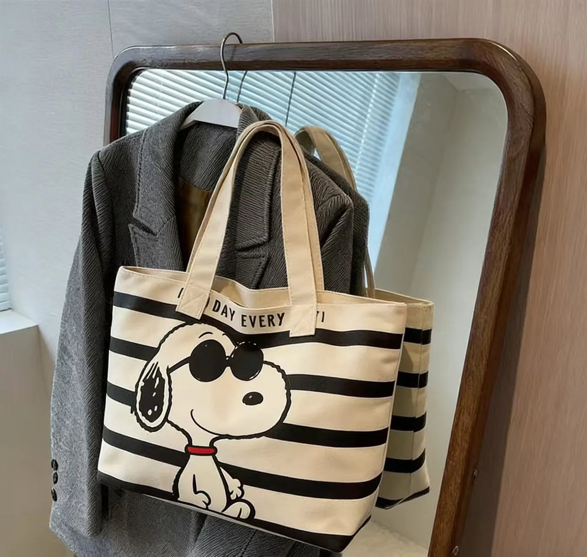 A beige and black striped canvas tote bag featuring a Snoopy cartoon face design, displayed on a hanger in front of a mirror.