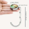 A silver bag hook with a round charm featuring colorful birds and flowers is shown against a hand.