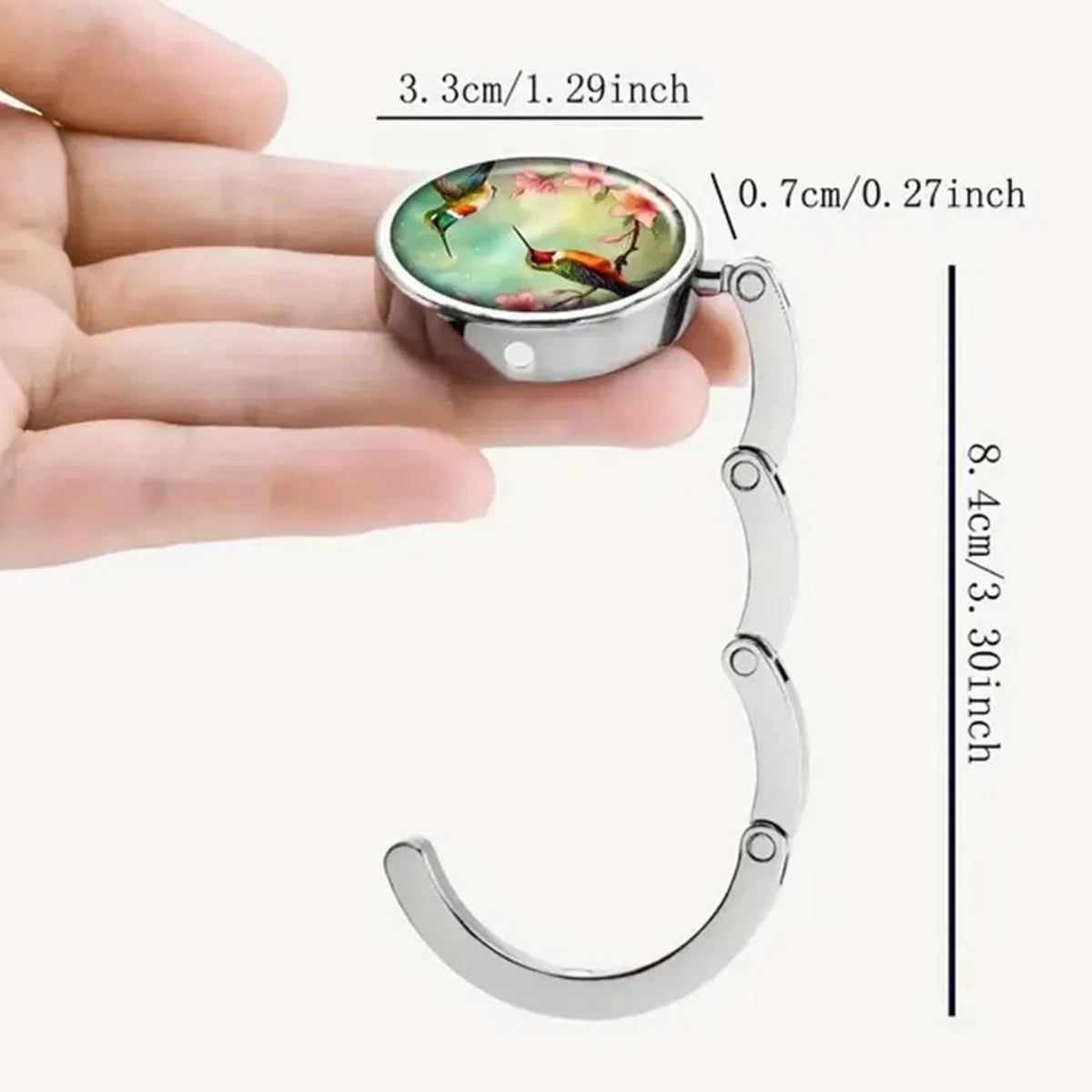 A silver bag hook with a round charm featuring colorful birds and flowers is shown against a hand.