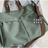 A light sage green nylon tote bag with brown straps and multiple internal pockets shown against a denim background.