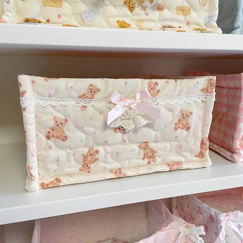 A cream quilted cotton storage basket with a pink lace flower design and a pink bow on a white shelf.