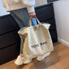 A person is holding a beige canvas tote bag with the text TOTE BAG in front of a dark wooden cabinet.