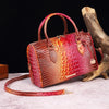 A reddish-brown crocodile pattern leather crossbody tote bag resting on a light surface next to a piece of driftwood.