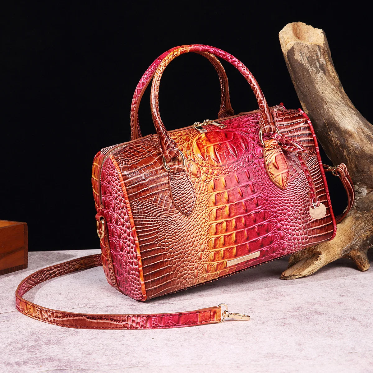 A reddish-brown crocodile pattern leather crossbody tote bag resting on a light surface next to a piece of driftwood.
