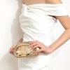 A woman holds an elegant gold-tone evening clutch bag against a white dress.