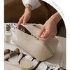 A beige foldable cosmetic storage bag is being held by a hand, next to a glass bottle and a makeup brush.
