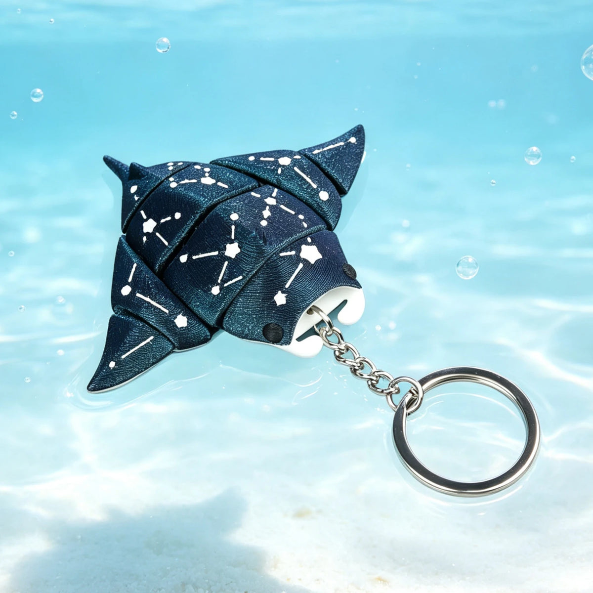 A dark blue fabric devil ray figurine with a silver chain attached, floating in clear water.