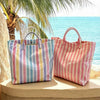 A pink and blue striped waterproof PVC shoulder tote bag resting on a ledge near the ocean with palm fronds in the background