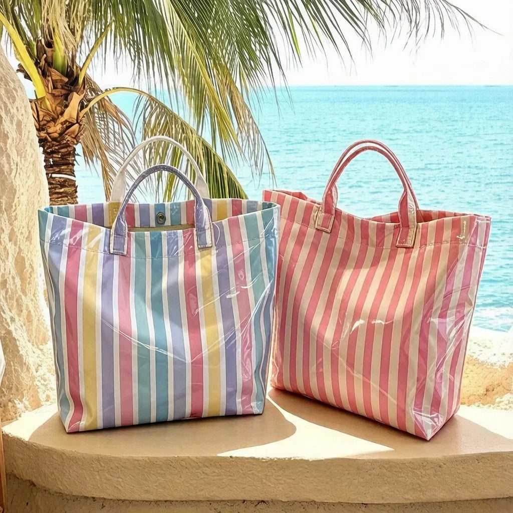 A pink and blue striped waterproof PVC shoulder tote bag resting on a ledge near the ocean with palm fronds in the background