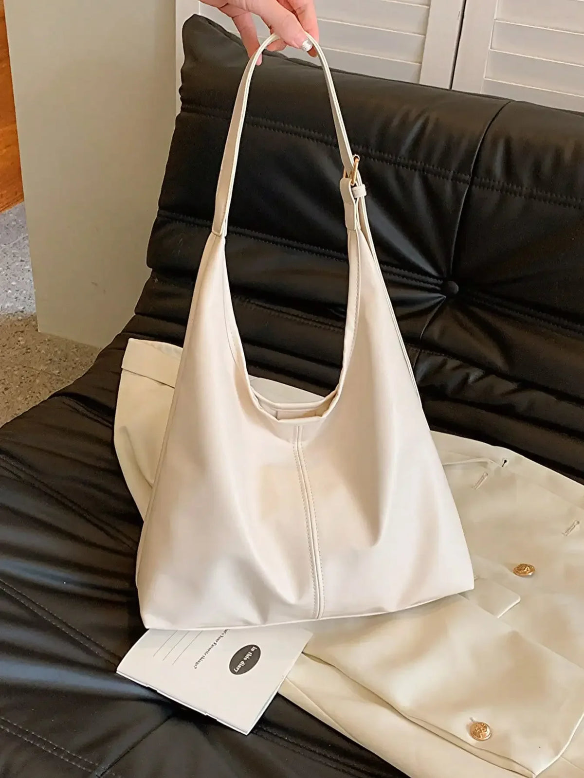 A light beige PU leather slouchy hobo shoulder bag is being held over a black leather couch.