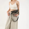 Eine Frau in weißem Crop-Top und beigen Hosen hält eine schwarze Leopardenmuster-Crossbody-Tasche.
