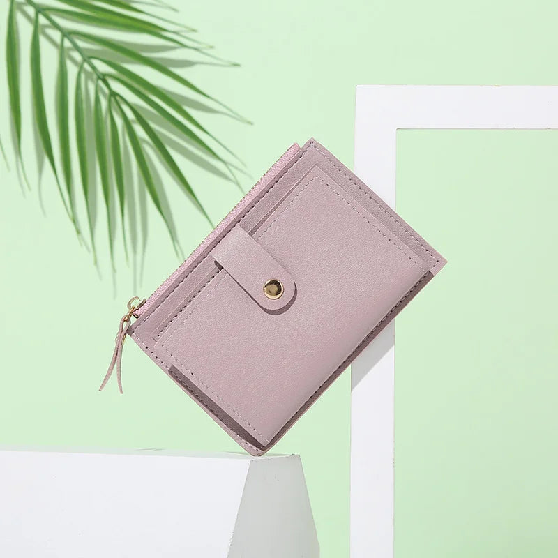 A light pink PU leather mini card holder wallet resting on a white surface with green foliage in the background.