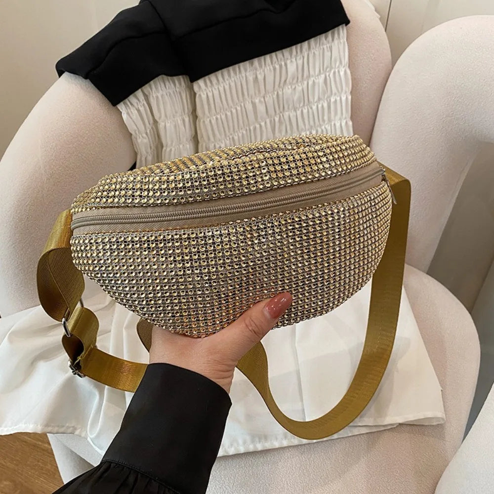 A hand holds a gold-toned, rhinestone-embellished beige crossbody waist bag with a matching strap.