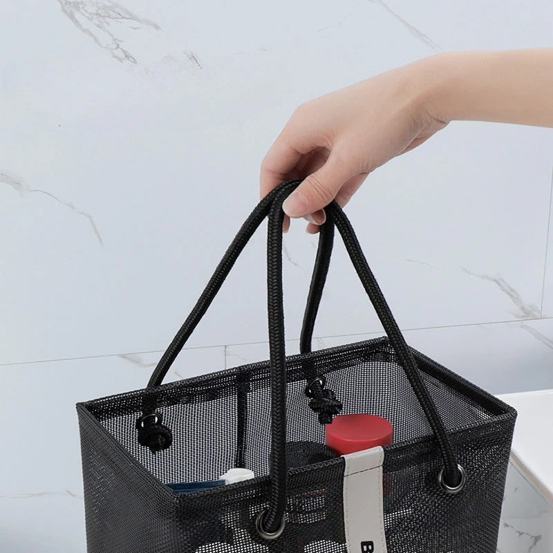 A hand is holding a black mesh drainage shower tote with sturdy handles against a white marble background.