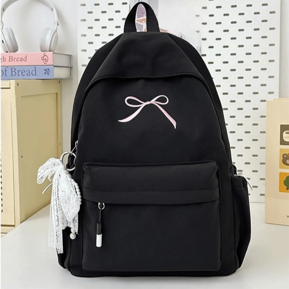 A black portable embroidered backpack with a white bow and decorative tassels is shown on a white surface.
