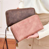 A woman holds a brown and pink double-zipper wallet with a leather strap against a light beige blazer.