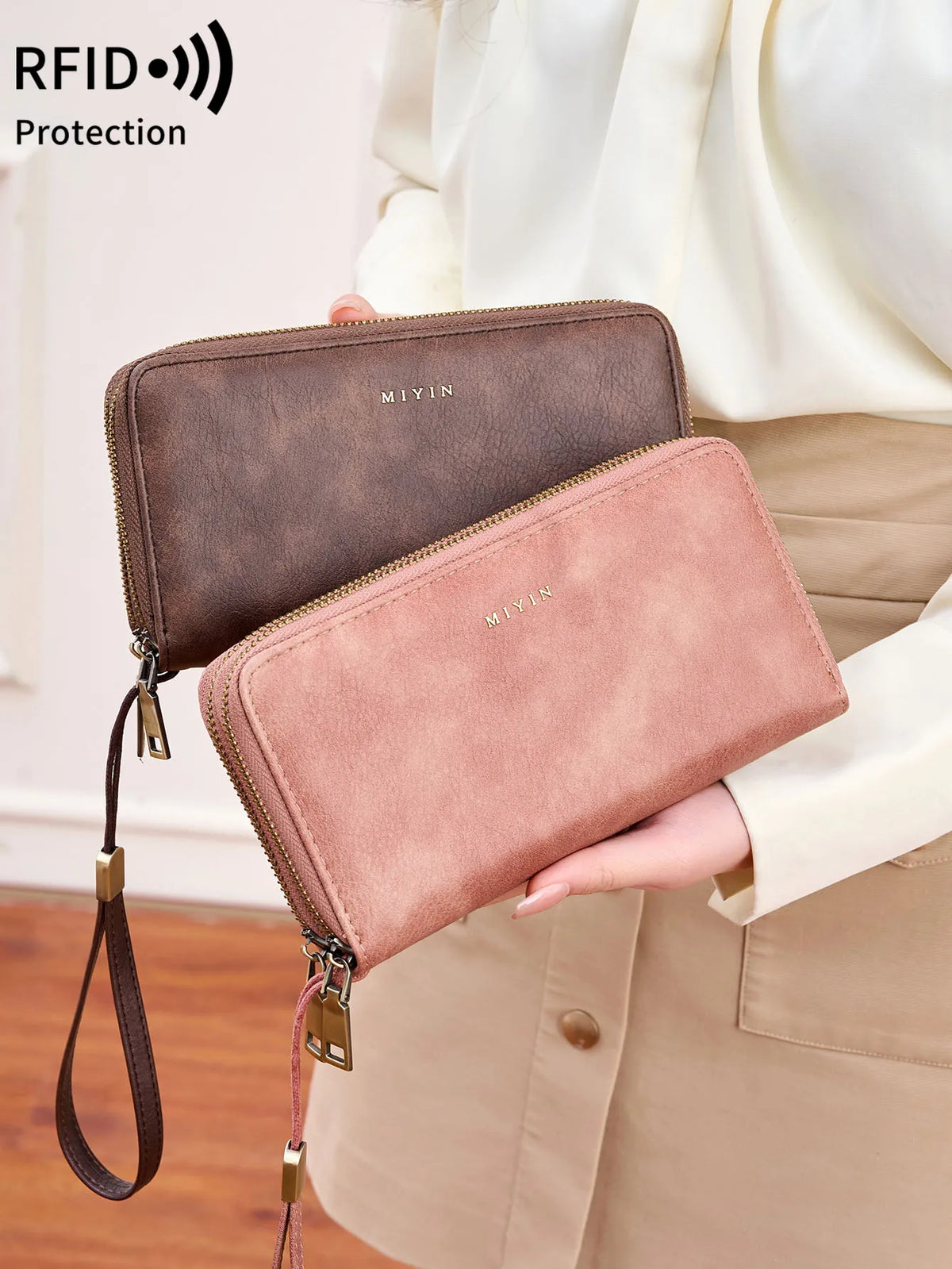A woman holds a brown and pink double-zipper wallet with a leather strap against a light beige blazer.