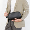 Un homme tient un sac pochette en cuir PU noir avec une sangle devant un blazer beige clair et un pantalon gris.