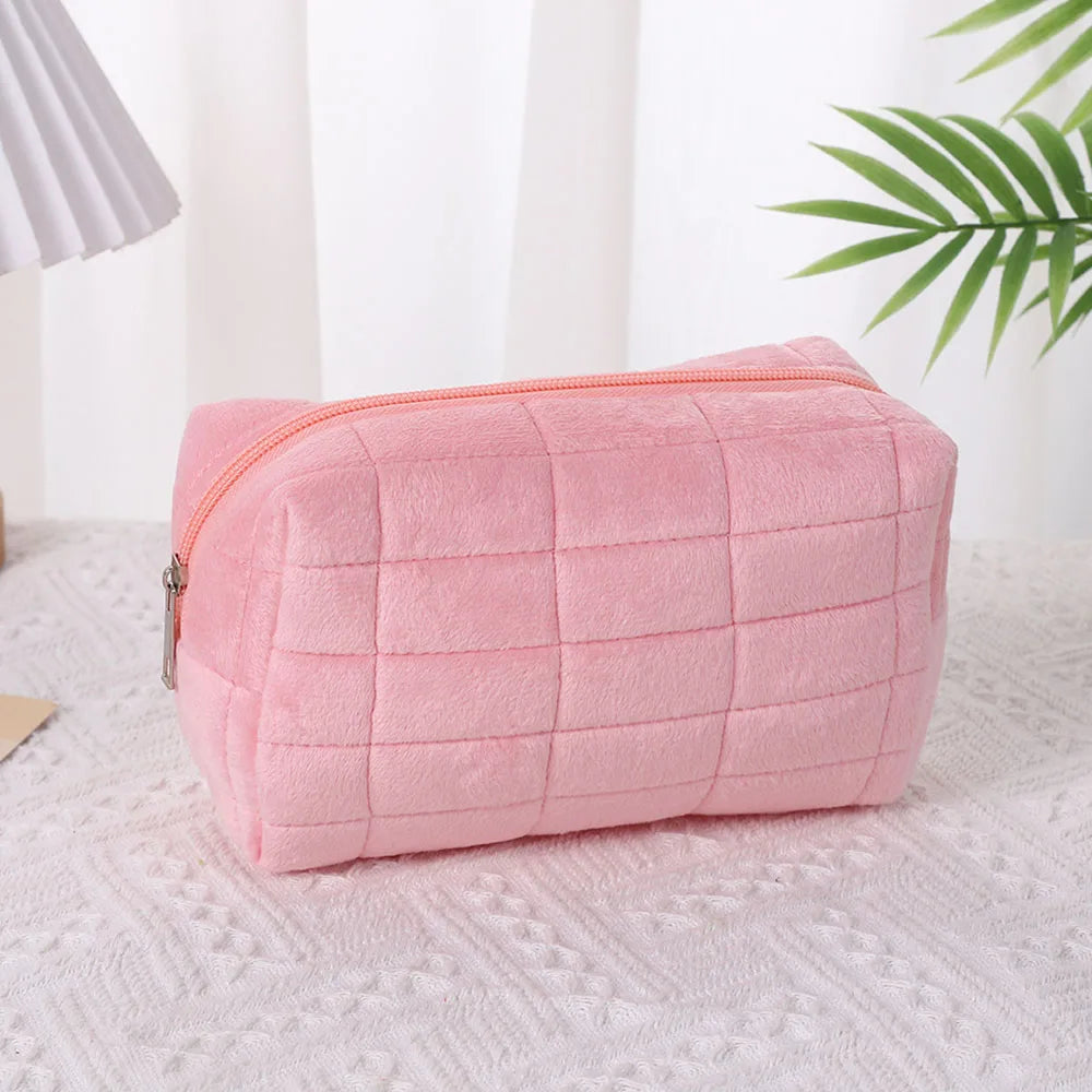 A pink quilted velvet cosmetic and toiletry bag resting on a white surface next to a green plant.