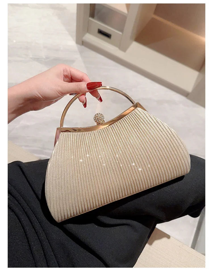 A hand holds a beige pleated glitter evening handbag with a gold top handle and chain strap against a dark background.