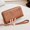 A brown leather wallet with a zipper closure and strap is held in a person's hand on a light surface.