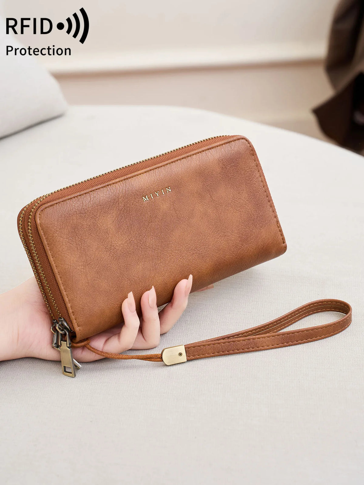 A brown leather wallet with a zipper closure and strap is held in a person's hand on a light surface.