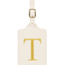 Etiqueta de equipaje rectangular de cuero color crema con una T bordada en oro y un clip metálico plateado.