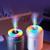 A white humidifier device emitting a purple mist, placed on a wooden desk next to a dark gray diffuser.