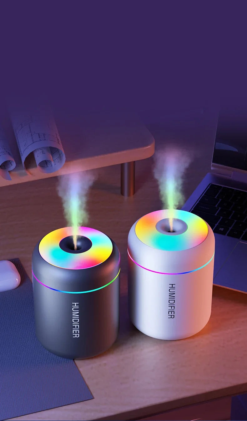 A white humidifier device emitting a purple mist, placed on a wooden desk next to a dark gray diffuser.