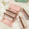 A pink portable detachable makeup bag with a beige strap and clear cosmetic pouches resting on white fluffy material.