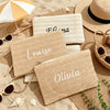 Three handwoven straw makeup bags with names Flora, Louise, and Olivia resting on sand.