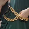 Primer plano de una correa de bolso de acrílico dorado llevada en la muñeca contra un tejido verde oscuro.