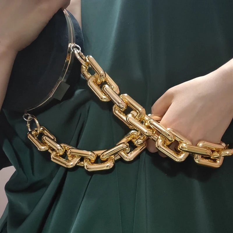 Primer plano de una correa de bolso de acrílico dorado llevada en la muñeca contra un tejido verde oscuro.