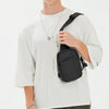 Modelo masculino vistiendo camiseta blanca y pantalones oliva, sosteniendo una bolsa cruz multifuncional negra.