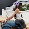 A woman wearing a white tank top and jeans sits on a wooden bench outdoors holding a black leather tote bag.