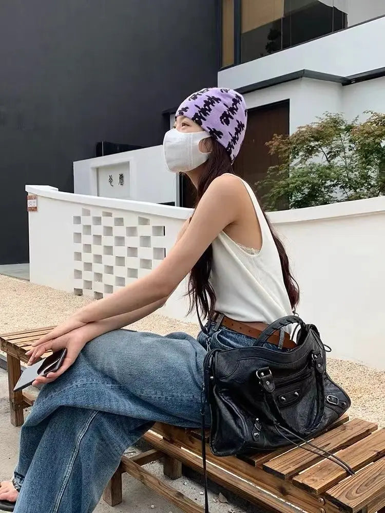 A woman wearing a white tank top and jeans sits on a wooden bench outdoors holding a black leather tote bag.