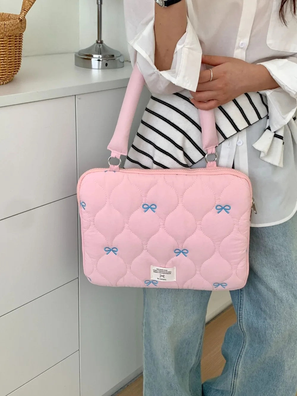 Bolso de portátil acolchado rosa con detalles de lazo azul, sostenido por una persona con camisa blanca y jeans.
