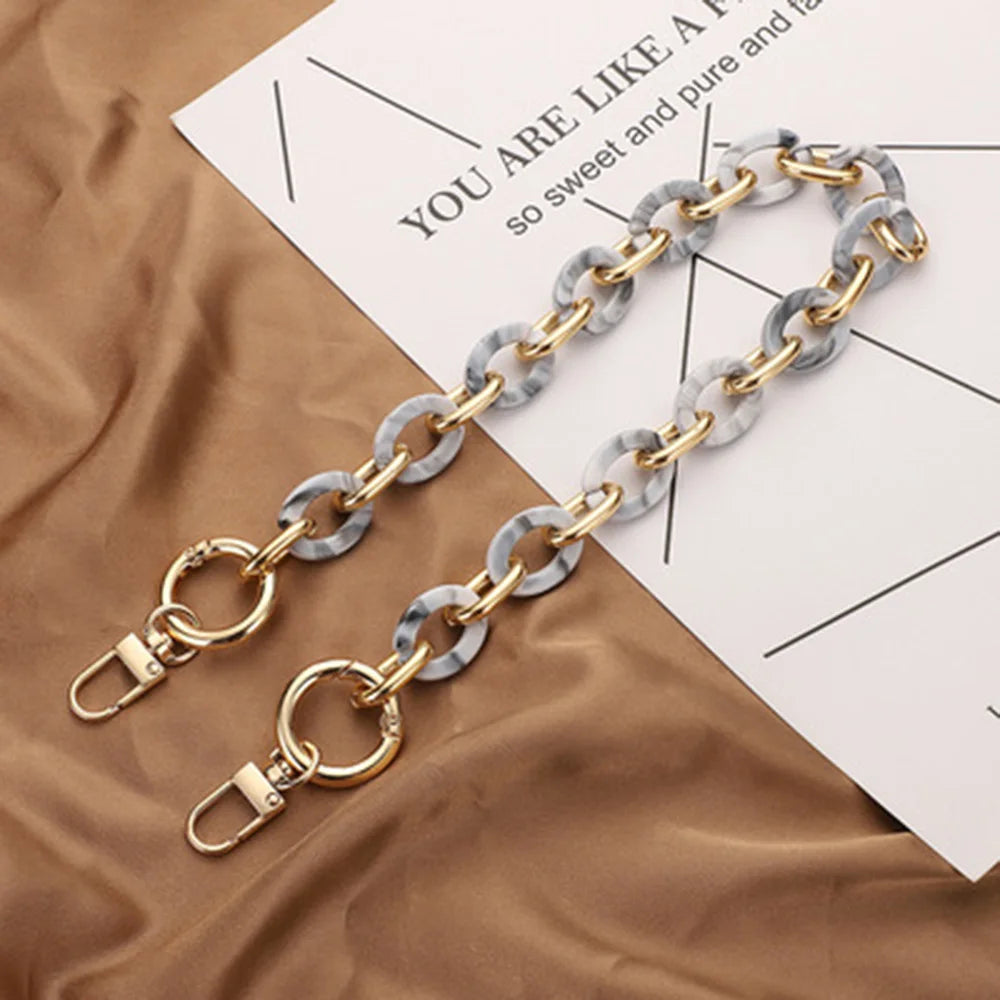 A pair of gold and grey marbled acrylic hand bag strap chains lying on a brown fabric next to a white paper.