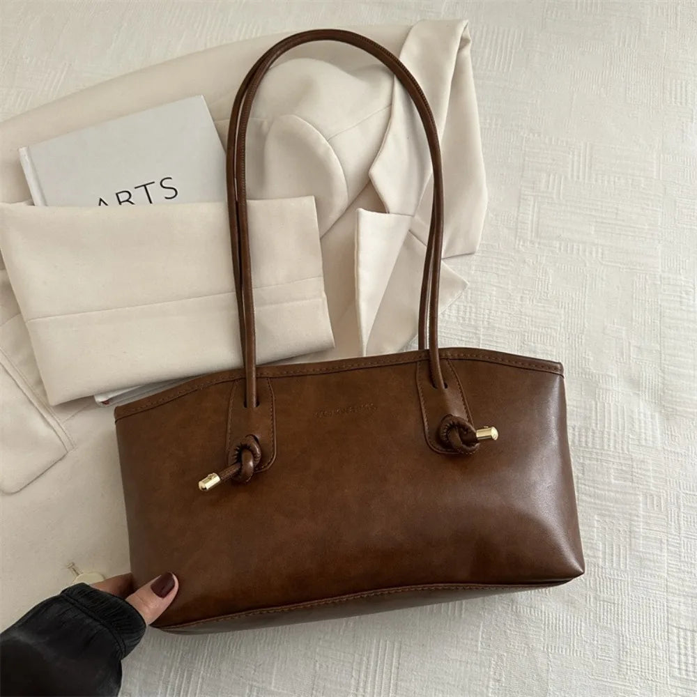 A brown leather tote bag with a handle is displayed on a white textured surface next to folded cream-colored paper.