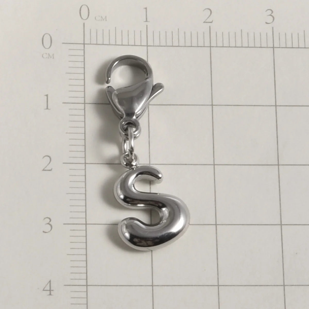 Silver stainless steel alphabet letter charm shaped like the letter S with a lobster clasp, measured against a ruler.