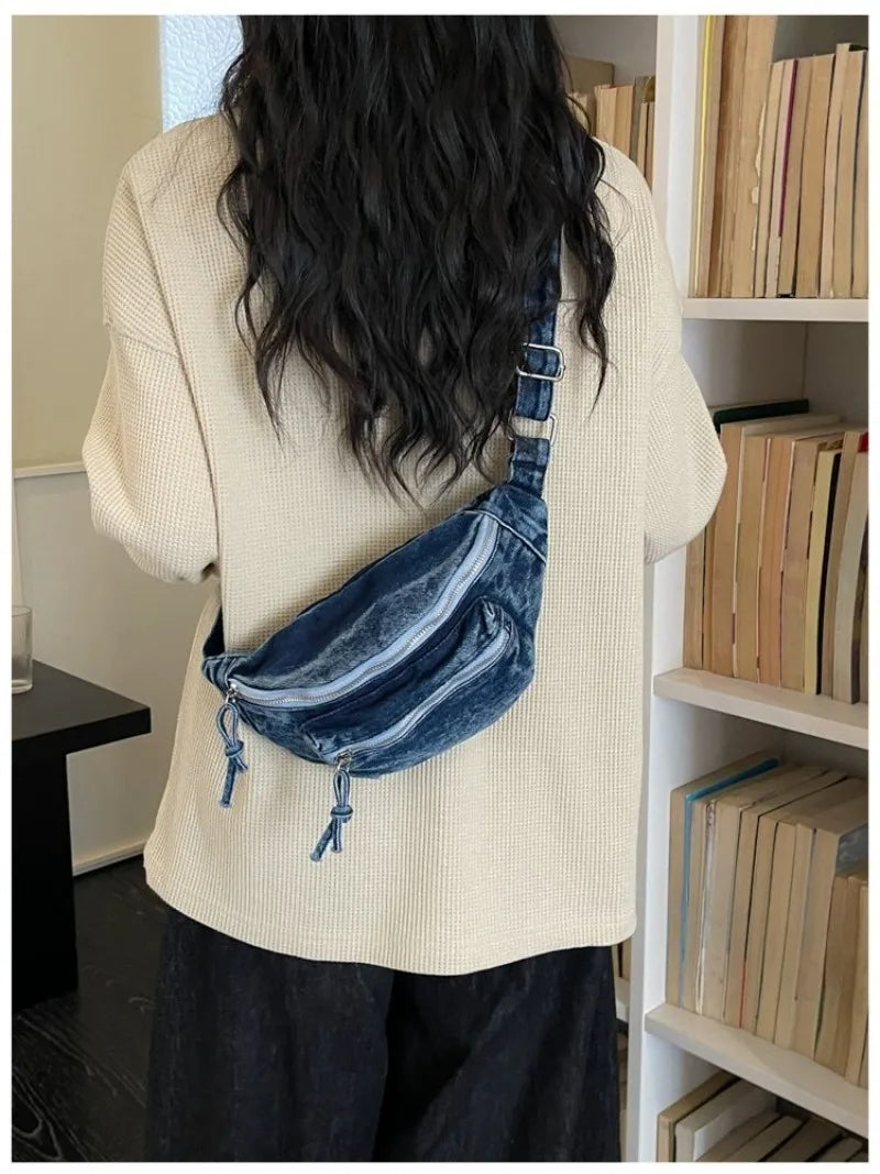 Une personne portant un pull tricoté crème et un pantalon foncé tient un sac bandoulière en denim délavé vintage.