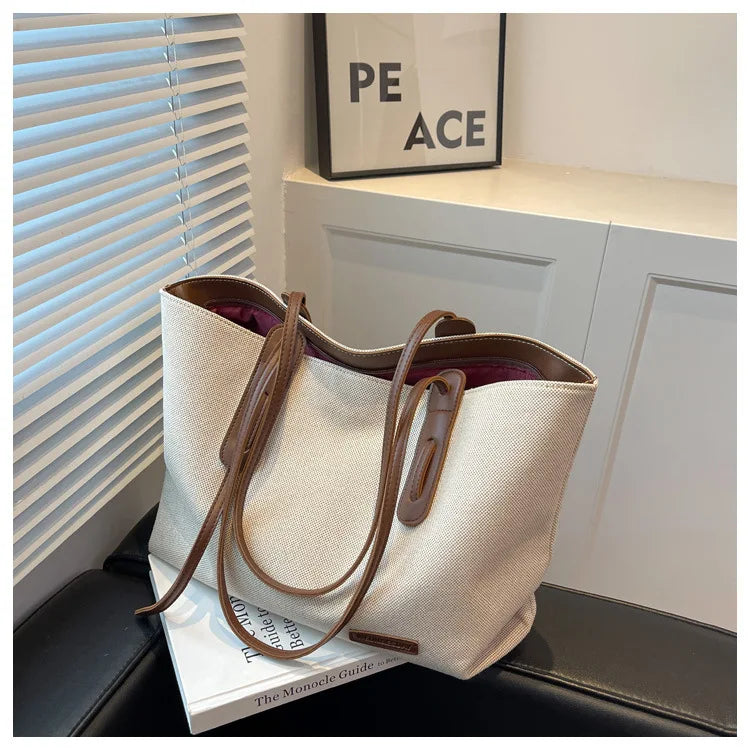 A beige canvas shoulder tote bag with brown leather straps resting on a black surface next to a white cabinet.