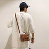 A man wearing a white t-shirt and a dark baseball cap is shown from the back, carrying a brown leather crossbody bag.