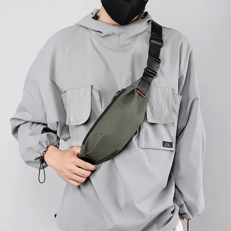 A person wearing a gray hooded jacket holds a dark olive green water-resistant nylon waist bag.
