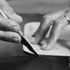 A close-up black and white photo shows a hand using a knife to cut leather for a leather artist.