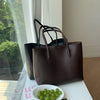 A dark brown vintage large-capacity tote bag with black handles is displayed on a white surface next to a bowl of green grape