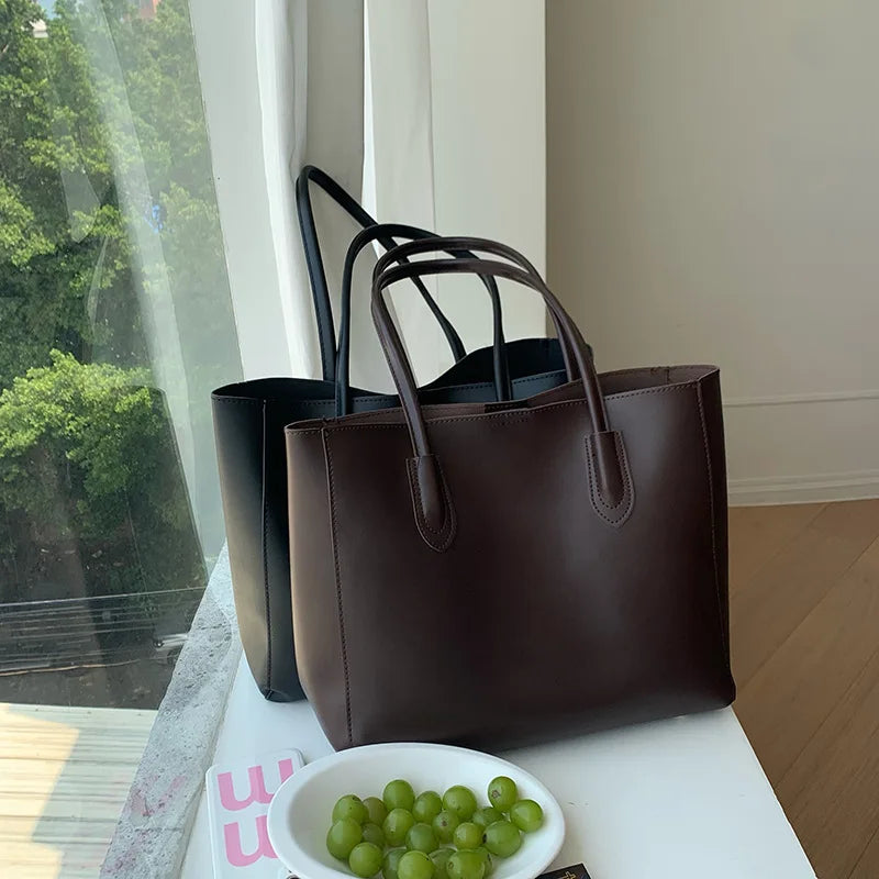 A dark brown vintage large-capacity tote bag with black handles is displayed on a white surface next to a bowl of green grape